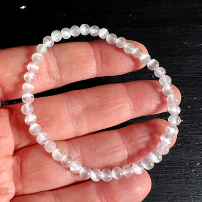 Selenit Armband aus kleinen milchig-weissen Perlen mit Katzenaugen-Glanz, gedehnt auf Fingern liegend | Happy Minerals